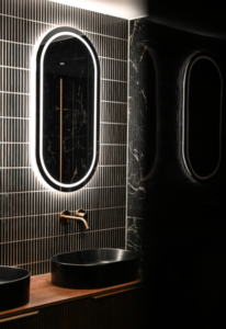Luxe badkamer in Putten met een ovale LED-spiegel, matzwarte opzetwastafel en een koperen inbouwkraan tegen een donkere tegelwand.