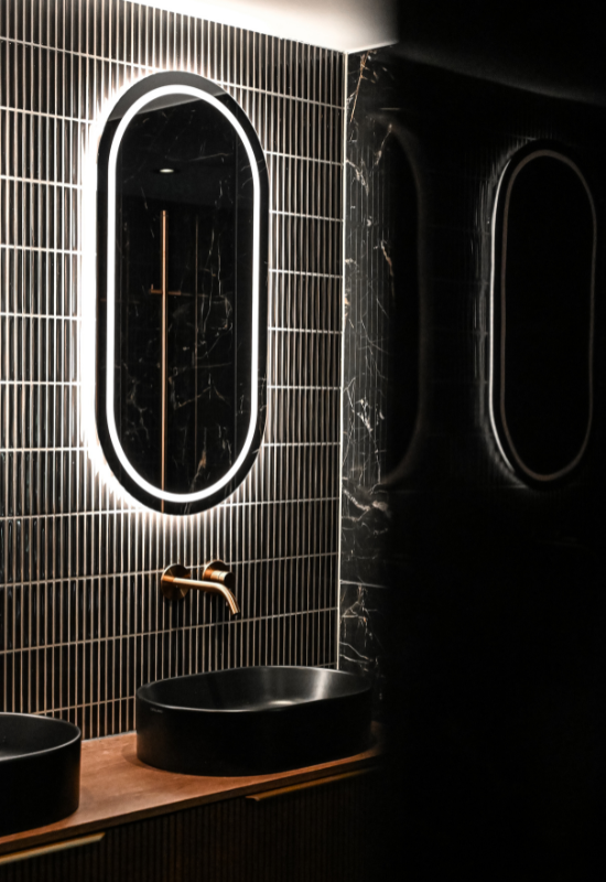 Luxe badkamer in Putten met een ovale LED-spiegel, matzwarte opzetwastafel en een koperen inbouwkraan tegen een donkere tegelwand.